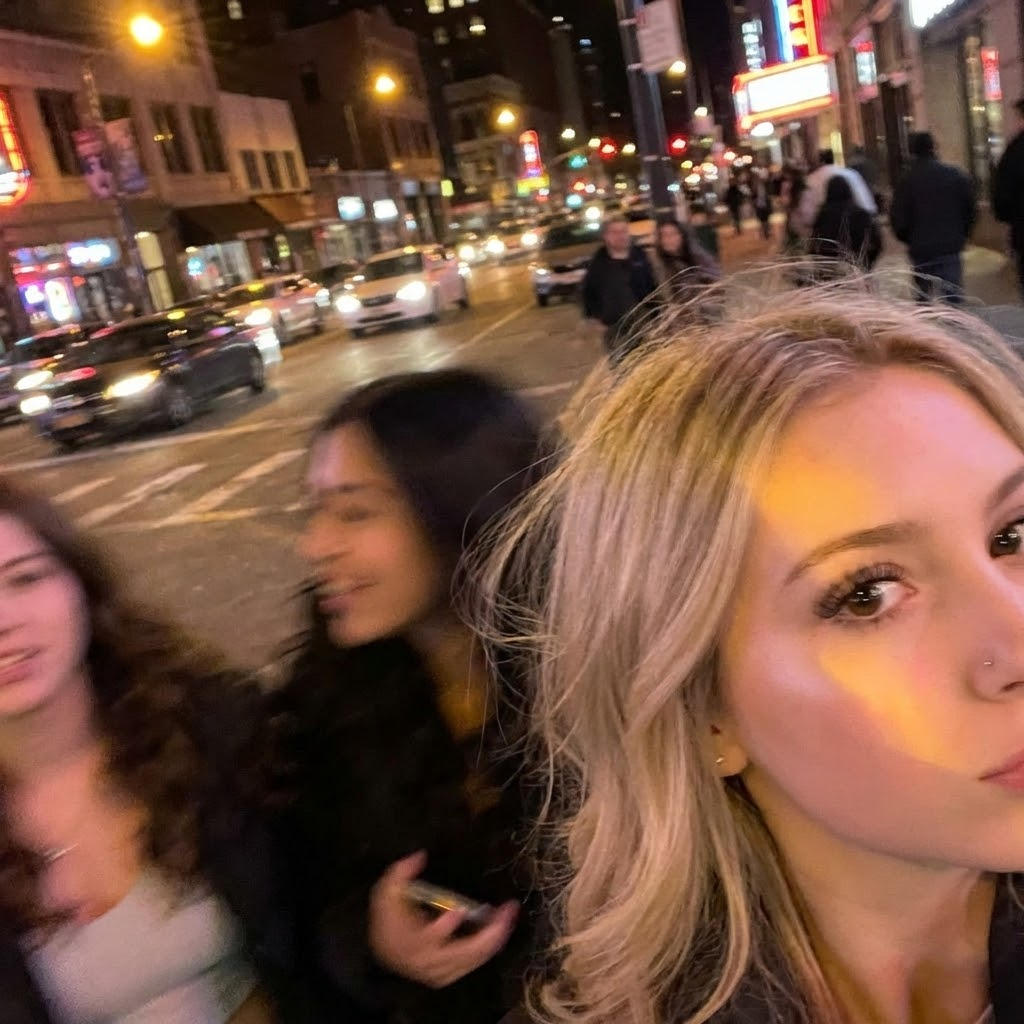 Take an extremely ordinary and unremarkable iPhone selfie, with no clear subject or sense of composition—just a quick accidental snapshot. The photo has slight motion blur and uneven lighting from streetlights or indoor lamps, causing mild overexposure in some areas. The angle is awkward and the framing is messy, giving the picture a deliberately mediocre feel, as if it was taken absentmindedly while pulling the phone from a pocket. The main character and the one taking the photo is the girl in the attached photo, next to her can be more people, this photo should display an imperfect moment. The background shows a lively TEMPAT at night, with neon lights, traffic, and blurry figures passing by. The overall look is intentionally plain and random, capturing the authentic vibe of a poorly composed, spontaneous iPhone selfie.