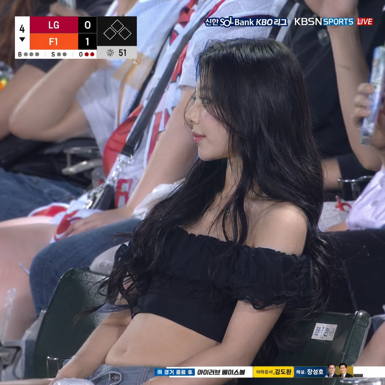 Create an ultra-realistic side view portrait of a candid KBO baseball broadcast screenshot of a woman accidentally caught on camera in the spectator seats. The team name is LG and F1. Her face should remain closer to the reference image than to a generic stadium fan. Do not change her into another person. Do not make her face wider, older, sharper, more westernized, or more idol-like. Keep the same delicate studio-portrait identity, but translated naturally into a real stadium environment. She is seated among a lively Korean baseball crowd, leaning back, not looking at the camera, looking out at the field, with extra long, straight and curly black hair cascading down both sides of her face, and wearing a black off-the-shoulder ruffle crop top revealing her soft, full rounded stomach. Use a realistic far-distance broadcast camera look: telephoto com, add broadcast HUD.