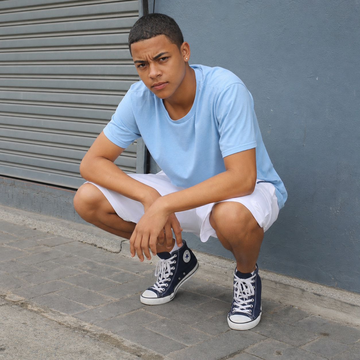 Young south african colored teen boy frowning short hair one ear pierced small gold stud light blue shirt white shorts navy blue hightop converse crouching on the pavement